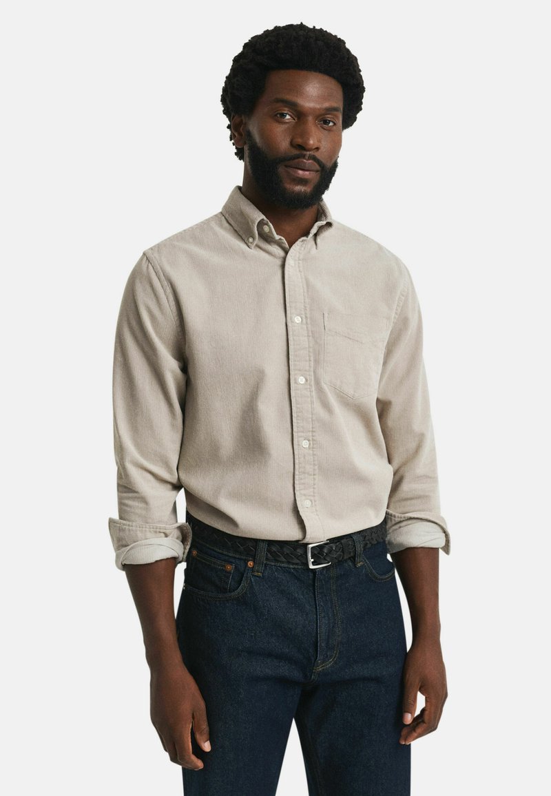 Hellbeige Langarmhemd mit einem Kentkragen, einer Brusttasche und aufgerollten Manschetten, kombiniert mit dunkelblauen Jeans und einem geflochtenen schwarzen Gürtel.