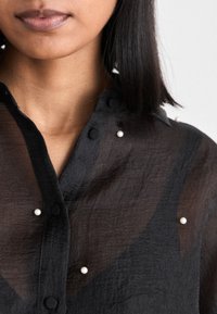 Gros plan d'une personne portant un chemisier noir transparent avec de petits boutons blancs en forme de perles et des cheveux foncés mi-longs visibles.