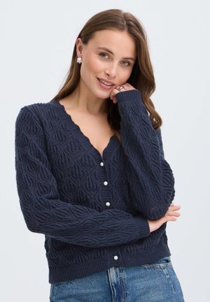 Femme aux cheveux châtain clair portant un cardigan texturé bleu marine avec des boutons en perle et un jean bleu, souriant avec la main près du visage.