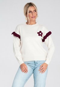 Blondynka ubrana w biały sweter z bordowym kwiatem i falbankami, jasnoniebieskie dżinsy i złote kolczyki w kształcie kół, pozująca z rękami na biodrach.