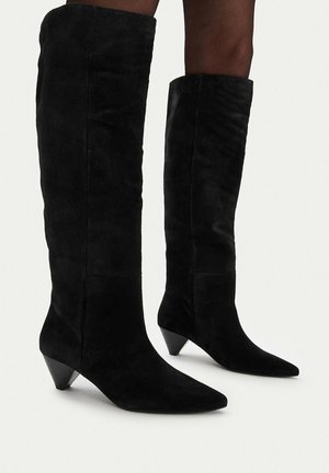 Botas negras de ante hasta la rodilla con punteras puntiagudas y tacones cónicos, usadas por una persona de piel oscura sobre un fondo liso.