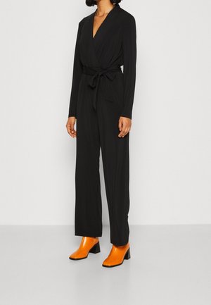 Kobieta ubrana w czarny kombinezon z długim rękawem zapinany na kopertę z wiązaniem w talii, do tego jasnopomarańczowe botki oraz złoty naszyjnik.