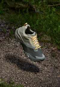 Lightweight gray Gore-Tex shoe with a textured black mesh section, yellow laces, and cushioned soles, elevated above a gravel surface.