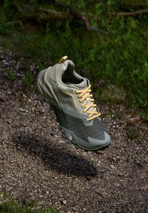 Kevyt harmaa Gore-Tex-kengät, joissa on teksturoitu musta verkkokappale, keltaiset nauhat ja pehmustetut pohjat, kohotettu sorapinnan ylle.