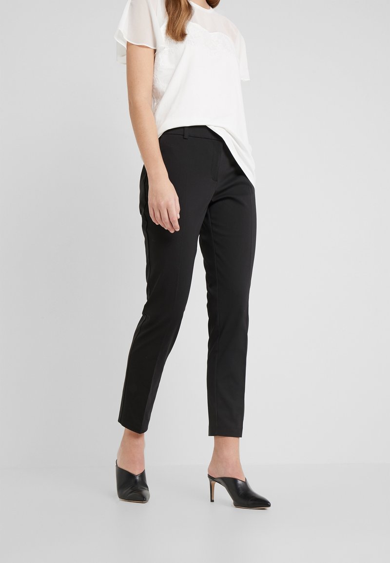 Schwarze, taillierte Hosen im Slim Fit mit glatter Textur und Seitentaschen. Kombiniert mit einem weißen, halbtransparenten Oberteil und schwarzen Mules.