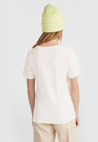 Lichtbeige katoenen T-shirt met korte mouwen en ronde hals, van de achterkant getoond, gecombineerd met een heldergroene beanie.