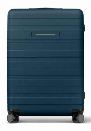 Valise rigide bleu foncé avec des rainures horizontales, une poignée supérieure et quatre roues pivotantes noires sur fond blanc.