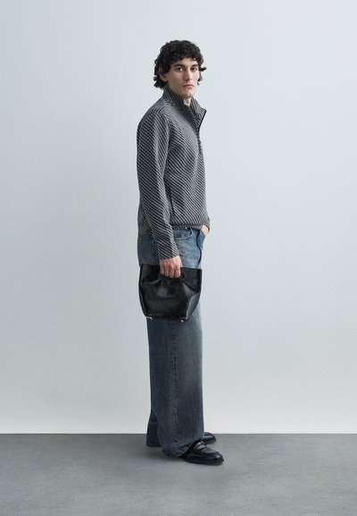 Sivý pruhovaný pulóver so štvrťzipsom, svetlomodré džíny so širokými nohavicami, čierna kožená kabelka a čierne topánky, stojace na svetlom pozadí.
