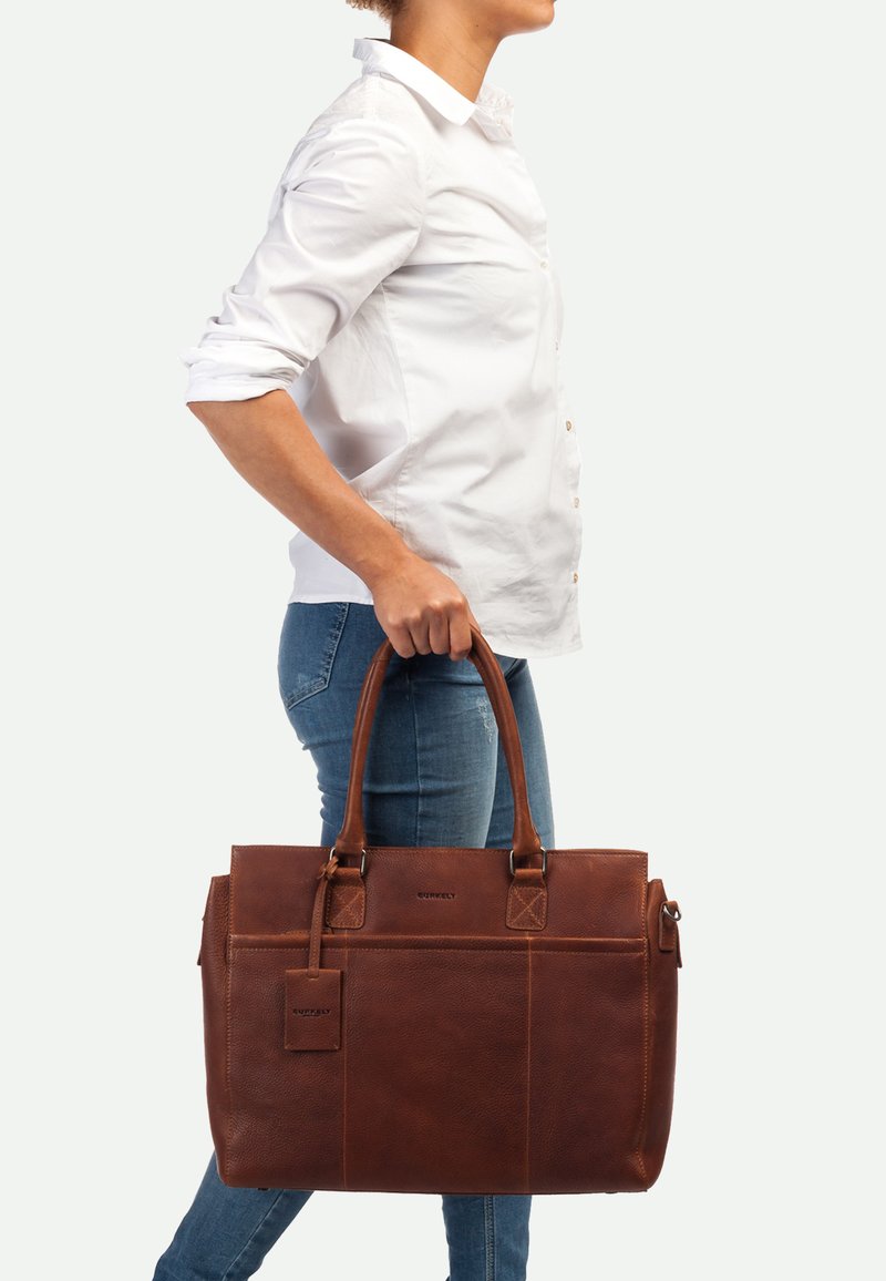 Bolso tote de cuero marrón con doble asas superiores, diseño estructurado y bolsillo exterior. Incluye una etiqueta de equipaje y acabado texturizado.
