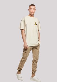 Beige crew-neck t-shirt with short sleeves, featuring a small yellow graphic on the chest. Paired with light brown cargo pants and white sneakers.