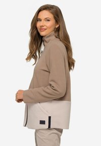 Fleecejacka med hög krage, med en beige övre del och en krämfärgad nedre del, dekorerad med en sidficka och svart logotyp.