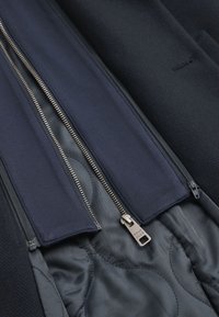 Marinblå rock med dubbla dragkedjor och silverfärgad detaljering. Inuti finns ett quiltat foder i en kontrasterande grå nyans som förbättrar textur och detalj.