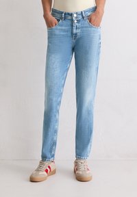 Persoon in lichtblauwe slim-fit jeans met opgerolde pijpen en beige sneakers met rode strepen, met de handen in de zakken, staande op een witte vloer.