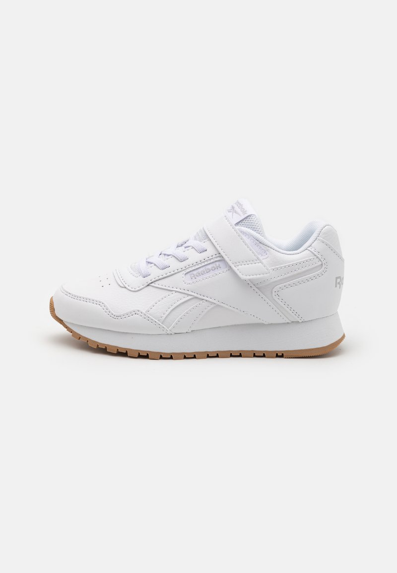 Baskets de sport blanches avec une tige en cuir lisse, un système de fermeture à lacets et une sangle Velcro, ainsi qu'une semelle en caoutchouc couleur gomme. Design simple avec des accents minimaux.