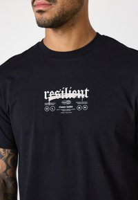 Camiseta de algodón negra con diseño gráfico blanco que incluye la palabra "resiliente" y detalles de la marca, cuello redondo y mangas cortas.