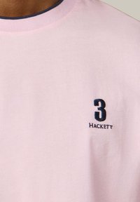 Maglietta rosa chiaro con scollo a girocollo, rifiniture blu navy, numero 3 ricamato in blu navy e piccolo logo "Hackett" sul petto.