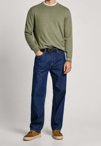 Olivgrüner Pullover mit Rundhalsausschnitt, kombiniert mit blauen geraden Jeans. Braune Sneakers mit hellen Sohlen. Schwarzer Gürtel mit einer schlichten Schnalle.