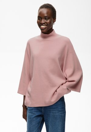 Femme souriante portant un pull rose ample à col montant avec des manches trois-quarts et un jean bleu, debout avec une main dans la poche.