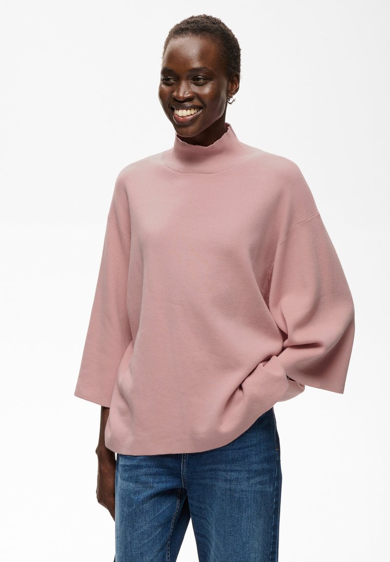 Femme souriante portant un pull rose ample à col montant avec des manches trois-quarts et un jean bleu, debout avec une main dans la poche.