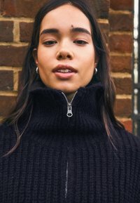 Crni pleteni džemper s visokim ovratnikom i zatvaračem na pola kopčanja. Ima rebrastu teksturu i srebrni detalj zatvarača.