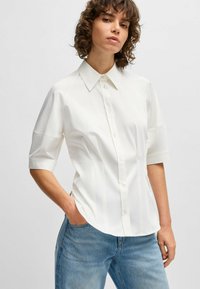 Camicia bianca a maniche corte con colletto a punta e pannelli laterali. Tessuto liscio con una leggera lucentezza; vestibilità sartoriale.