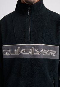 Sort fleece pullover med halv-lynlås, en grå stribe med hvid "Quiksilver" tekst og struktureret stof for varme og komfort.