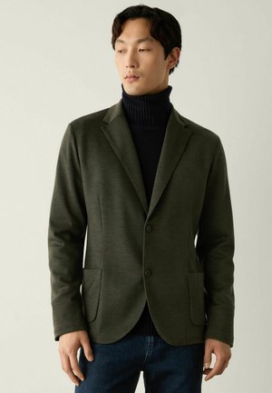 Mand iført en olivengrøn blazer over en sort rullekrave og blå jeans, ser lidt til siden mod en ensfarvet baggrund.