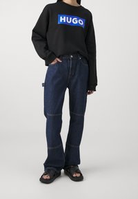 Black sweatshirt with a blue "HUGO" logo, paired with dark blue denim jeans featuring contrasting stitching and a side pocket. Black sandals.