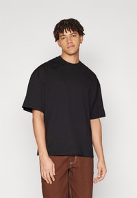 Sort bomulds T-shirt med afslappet pasform, rund halsudskæring og korte ærmer. Modellen har brune bukser på med hvide stikninger.