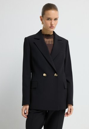 Zwarte dubbelgebreide blazer met geknoflookte revers, goudkleurige knopen en voorkruiszakken. Gedragen over een gestructureerde bruine top. Perfecte pasvorm.