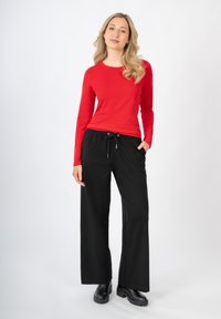 Nainen, jolla on pitkät vaaleat hiukset, pukeutuneena punaiseen pitkähiharaiseen paitaan, mustiin leveälahkeisiin housuihin, joissa on nauhavyö, ja mustiin nilkkureihin, seisoo valkoista taustaa vasten.