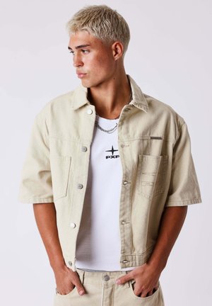 Joven con cabello rubio corto que lleva una chaqueta beige de manga corta y pantalones sobre una camisa blanca, mirando hacia su izquierda con las manos en los bolsillos.