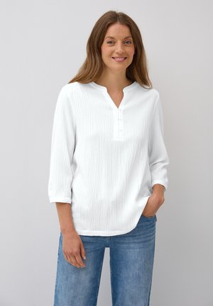 Blouse blanche texturée avec un col henley, comportant trois boutons, des manches trois-quarts et une coupe décontractée. Associée à un jean bleu.