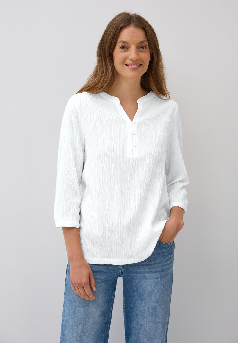White textured blouse with a henley neckline, featuring three buttons, three-quarter sleeves, and a relaxed fit. Paired with blue jeans.