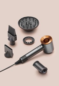 Haartrockner mit sechs abnehmbaren dunkelgrauen Styling-Aufsätzen, darunter Diffusor, Kamm und Konzentrator-Düsen, auf einer beigen Oberfläche ausgelegt.