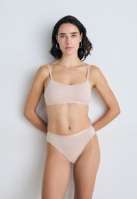 Vrouw met schouderlang donker haar, gekleed in een huidkleurige beha en ondergoed, staande tegen een effen witte achtergrond met handen achter de rug.