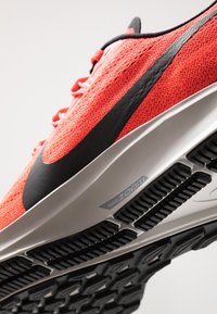 Gros plan d'une chaussure de course Nike Zoom corail montrant une tige texturée, le logo Swoosh noir et un motif détaillé de la semelle noire.