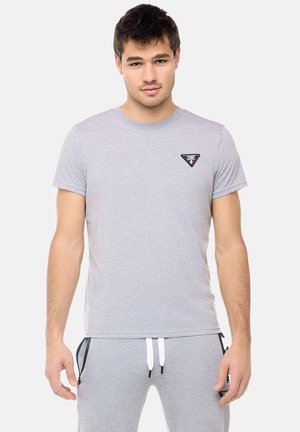 Jonge man draagt lichtgrijs t-shirt met klein borstlogo en bijpassende grijze joggingbroek met witte trekkoorden, staand tegen een witte achtergrond.