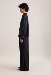 Person mit kurzen, lockigen Haaren, der im Profil steht, trägt eine marineblaue Satinbluse mit breiten Manschetten und schwarze, weit geschnittene Hose.