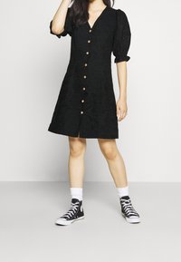 Vestito nero fino al ginocchio con colletto a V, maniche a sbuffo e chiusura con bottoni. Presenta una texture in pizzo e bottoni in legno. Abbinato a sneaker nere.