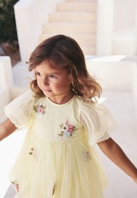 Gelbes Kleid mit transparenten Puffärmeln, verziert mit floraler Stickerei in Rosa, Blau und Grün. Weicher, fließender Tüllstoff und geraffte Taille.