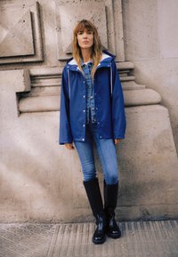 Ženska z dolgimi lasmi stoji ob teksturirani kamniti steni, oblečena v modro jakno, jeans srajco, kavbojke in črne škornje do kolen.