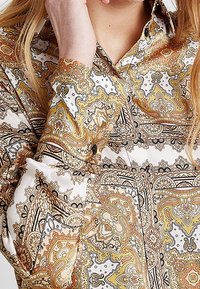 Långärmad skjorta med vit bas och intrikata paisleymönster i guld, orange och svart. Har en knapprad framtill och en slät tygtextur.