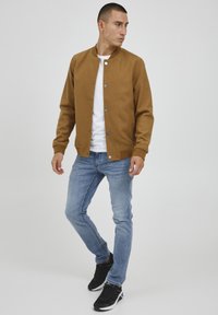 Chaqueta bomber marrón de un tejido suave con botones a presión, usada sobre una camisa blanca y combinada con jeans azul claro y zapatos negros.