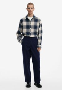 Kockovaná košeľa s gombíkmi v modrej a krémovej farbe, nosená na bielej okrúhlej krku, v kombinácii s tmavomodrými nohavicami a čiernou obuvou.