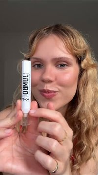 Hvid cylindrisk læbestiftbeholder med klar bund, mærket "NYX" og "Brow," holdt af en person med bølget hår, der fremviser dens design og branding.