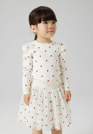 Jeune fille aux cheveux foncés portant une robe blanche ornée de petits cœurs rouges, debout devant un fond gris clair uni.