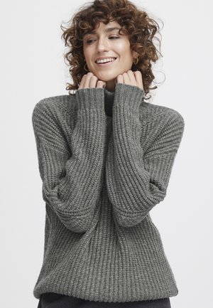 Kroeshaarige vrouw lacht, draagt een dikke grijze ribgebreide trui, handen dichtbij het gezicht, tegen een effen witte achtergrond.