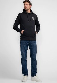 Čierna mikina s kapucňou, predným vreckom, grafikou na ľavej strane hrudníka a detailmi na rukávoch. Nosená s modrými džínami a bielymi teniskami.