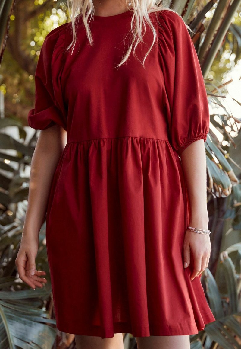 Robe en coton rouge avec manches courtes bouffantes, un col rond et une jupe froncée. Texture lisse, longueur au-dessus du genou.